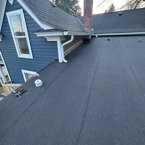 Finished low slope roof section next to a blue sided house and brick chimney